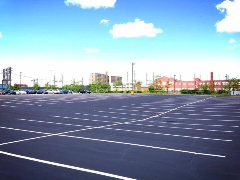 Empty parking lot with many lined spaces, cars parked in the distance, and buildings under a partly cloudy sky.