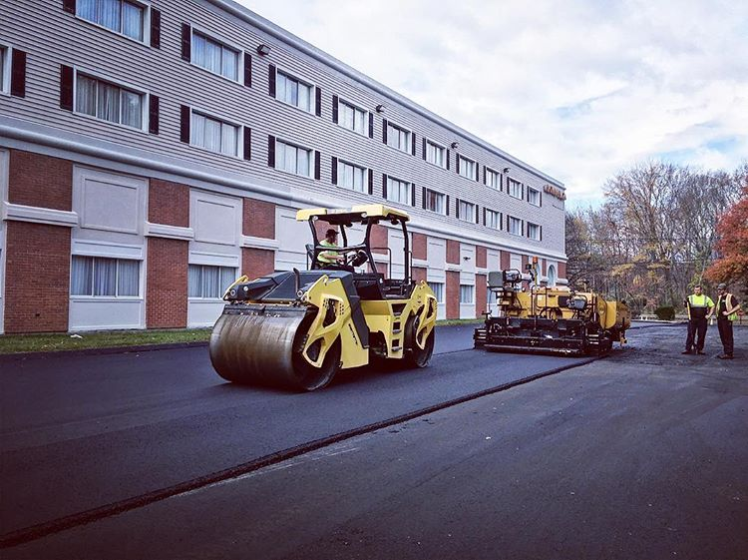 Construction workers operate a yellow road roller and a paving machine on a newly paved asphalt road in front of a multi-story brick and white hotel building during daytime.