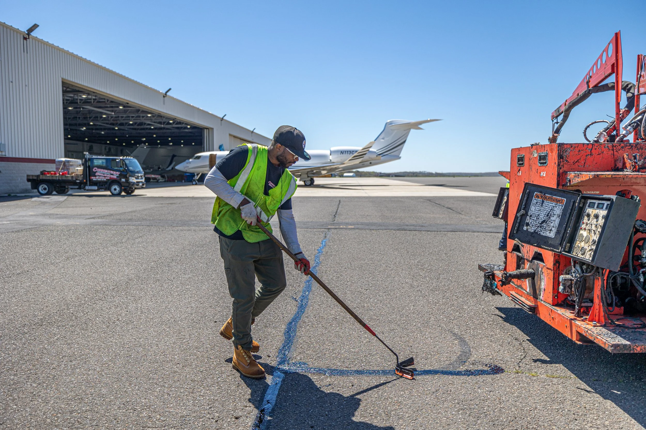 Man in a high-visibility vest and boots marking a line on the tarmac at an airport, with private jets and maintenance vehicles nearby.