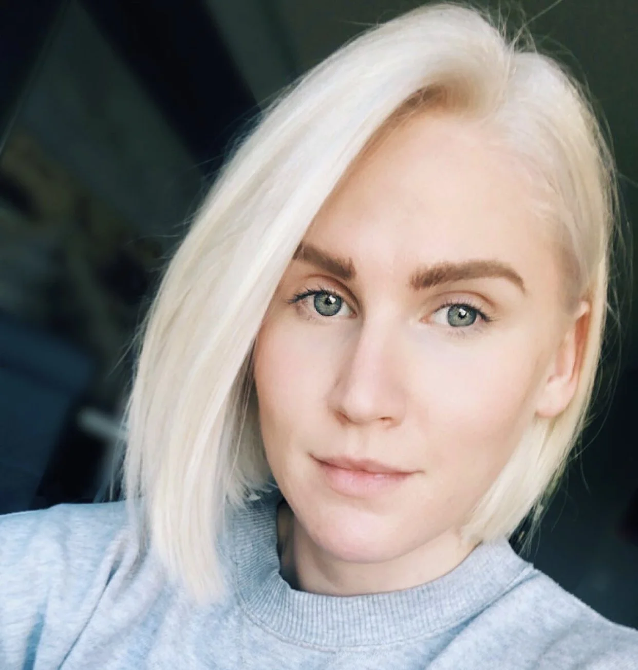 A young woman with platinum blonde hair, light eyes, and natural makeup, wearing a gray top, looking at the camera with a slight smile.