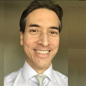 A smiling man in a white dress shirt and light-colored tie, taking a selfie indoors.