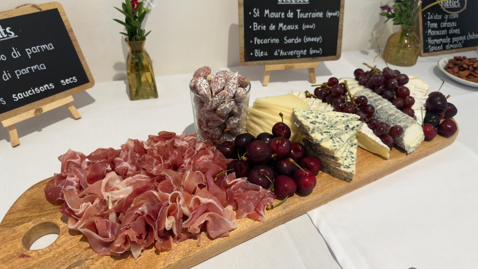 Assorted cheeses, cured meats, red grapes, and nuts on a wooden serving board at a cheese platter display.