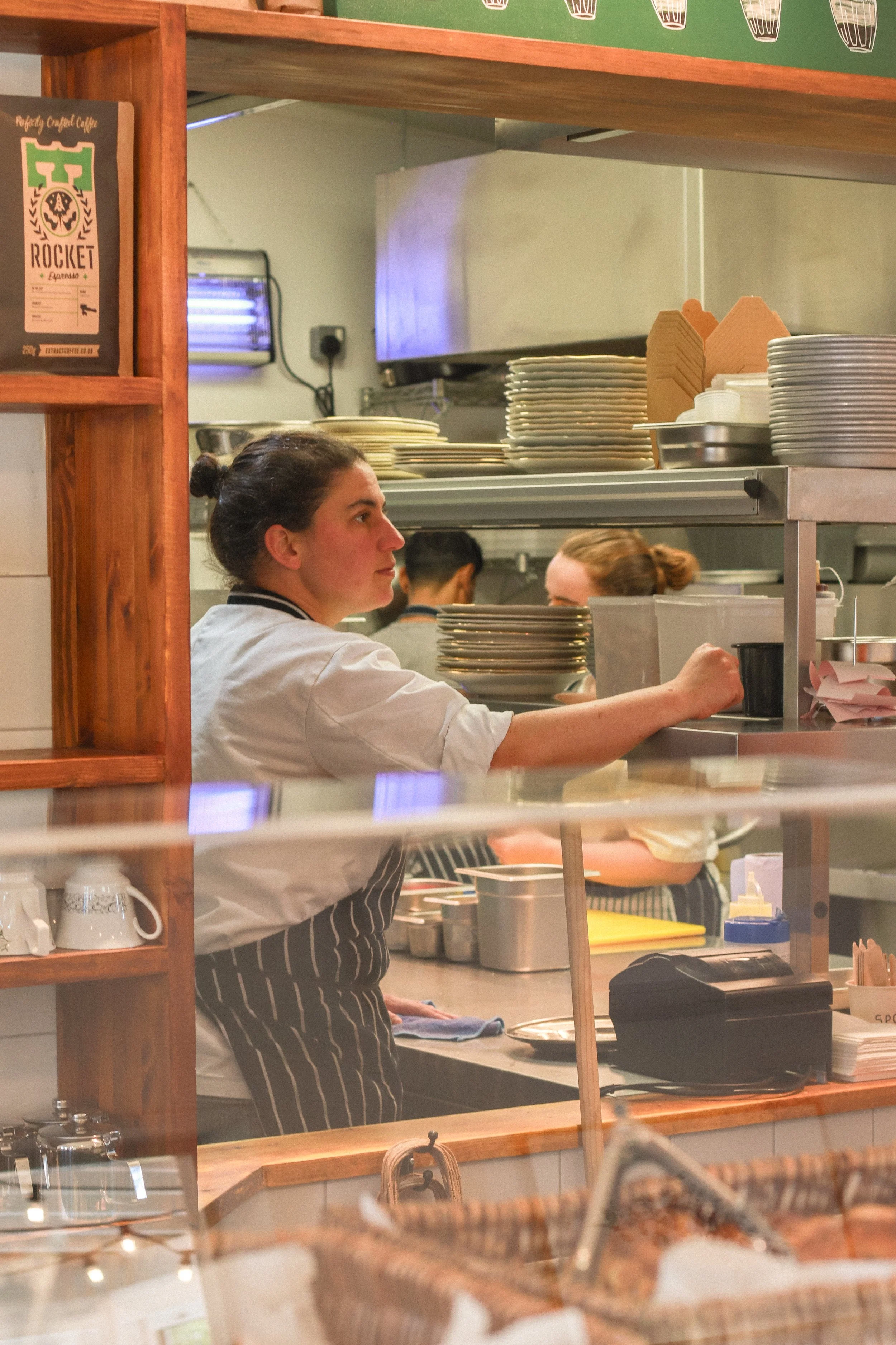 A female chef in a restaurant kitchen wearing a white uniform and black-and-white striped apron, working behind a serving counter with stacks of plates and kitchen utensils.