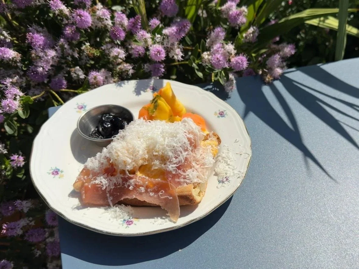 A plate with toasted bread with parmaesan cheese, with slice of Seranno ham, and a small dish of black olives on a blue table, with purple flowers and green foliage in the background.