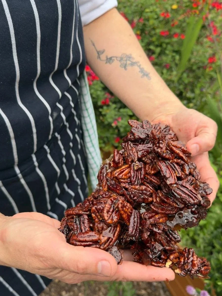 Person holding a large cluster of candied pecans outdoors, with a background of greenery and red flowers.