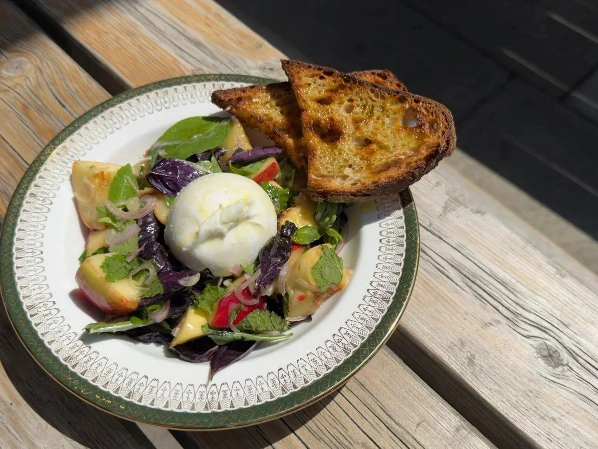 A salad with mixed greens, sliced peaches, fresh herb, topped with Burrata, served with two slices of toasted bread on a decorative plate.