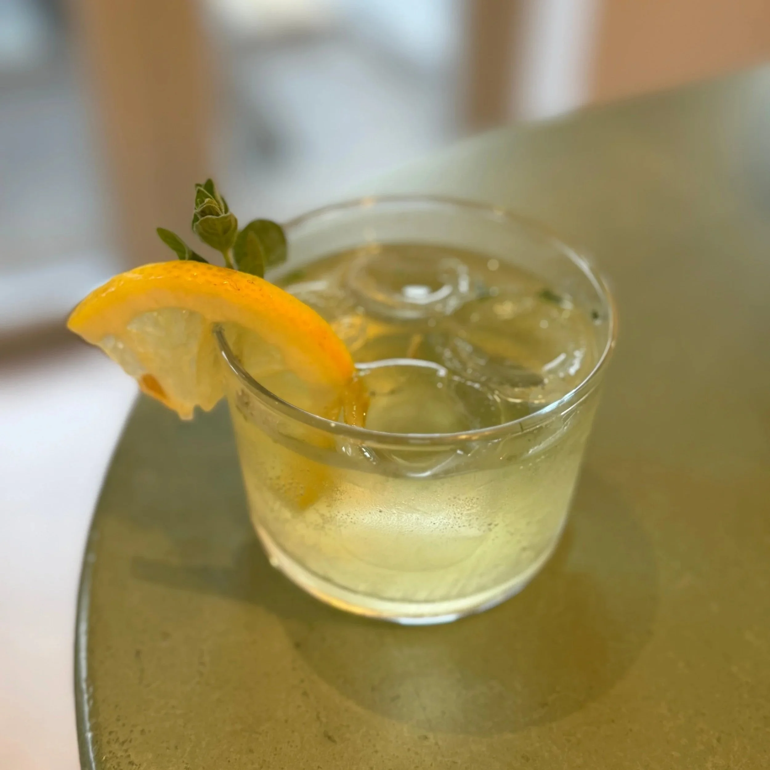 Close-up of a cocktail with a yellow lemon wedge and green leaves inside, viewed from below with a blurred background.