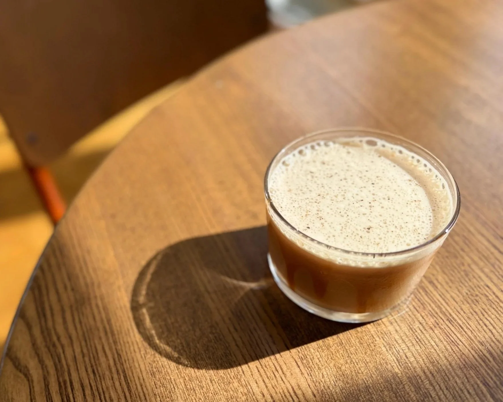 A clear glass of coffee cocktail with foam on top sitting on a wooden table.