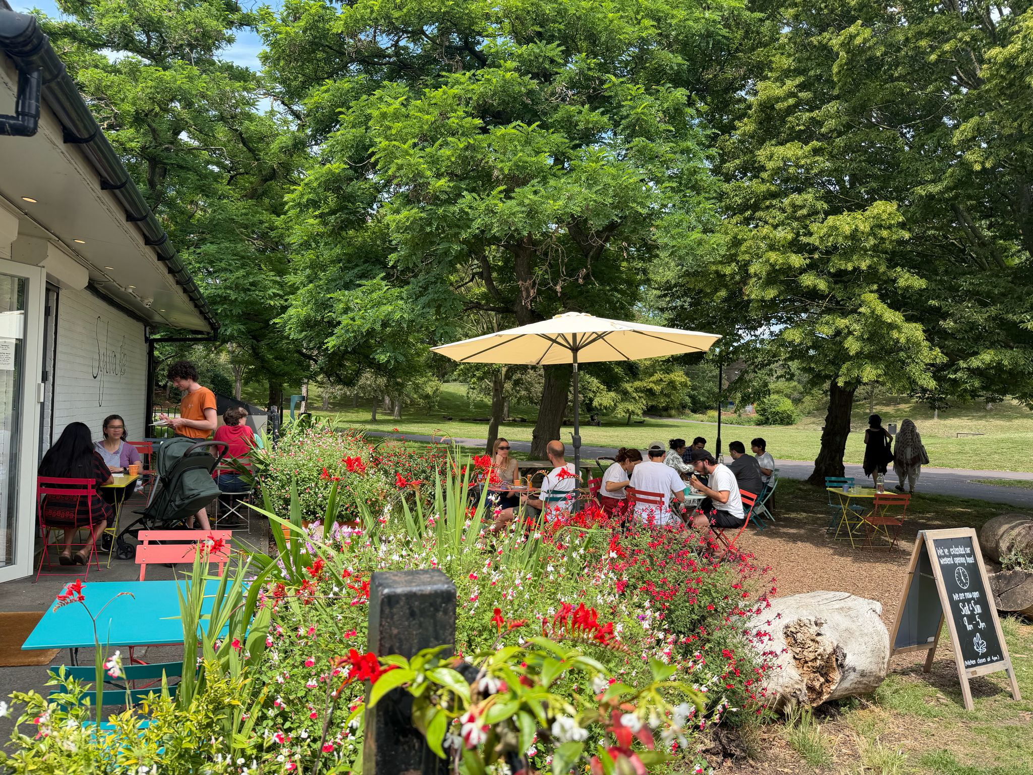 Outdoor patio area with tables, chairs, with a parasol, surrounded by colorful flowers and lush green trees, with people dining and socializing.