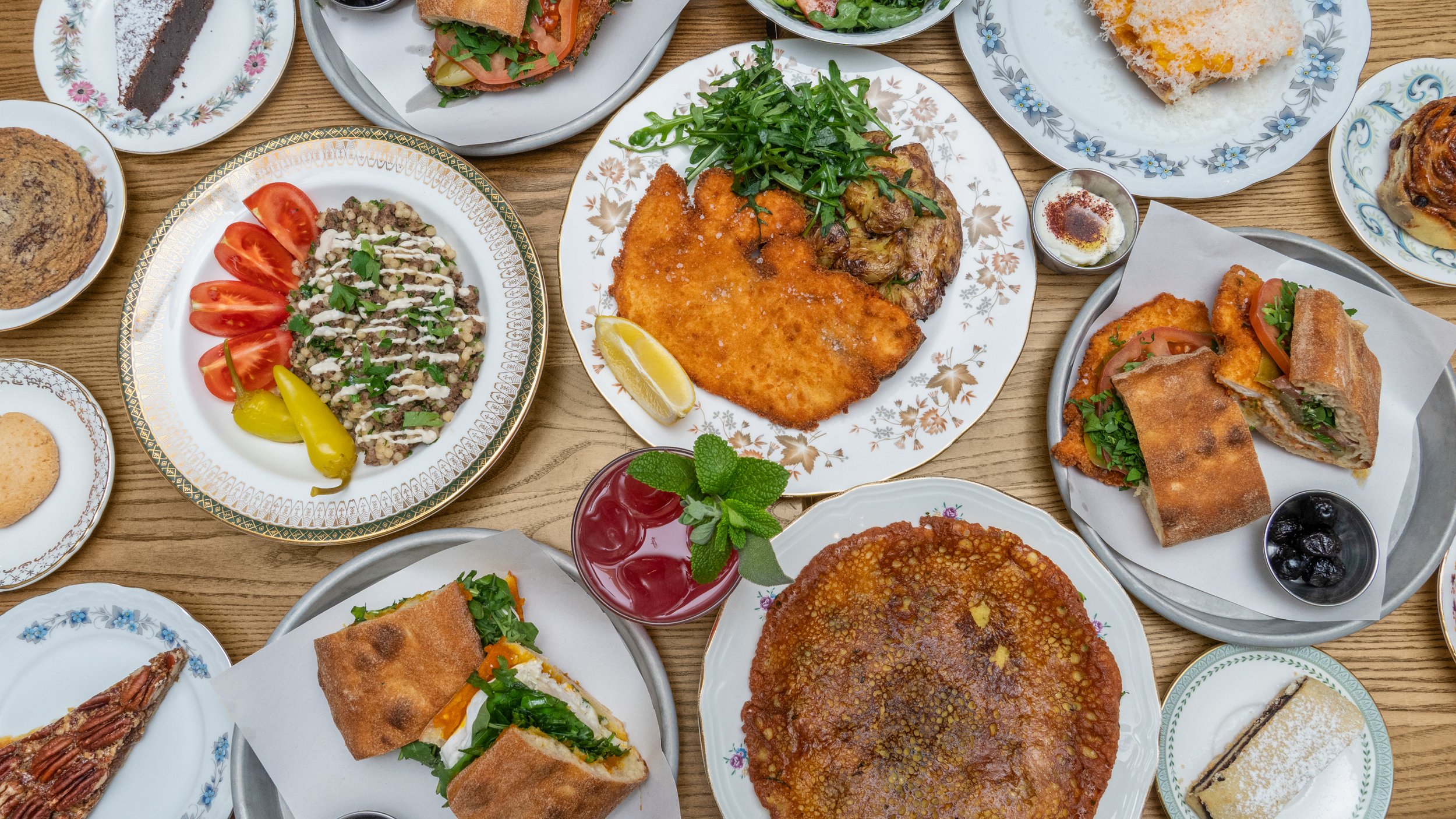 A table filled with various plates of food including salads, sandwiches,  a slice of cake, and olives, all on decorative dishes.