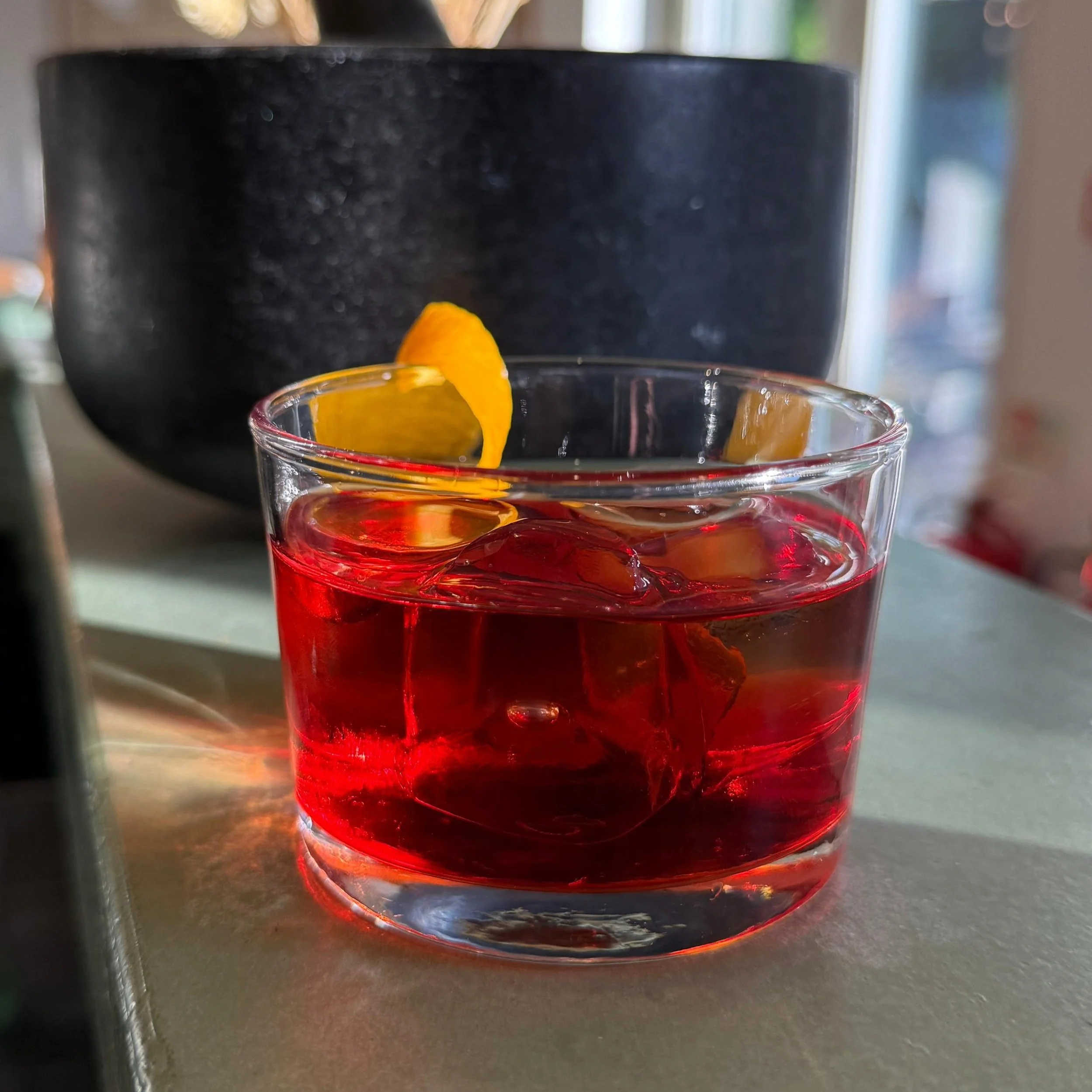 A glass  filled with a red beverage and ice cubes, garnished with an orange twist, placed on a light-colored surface.