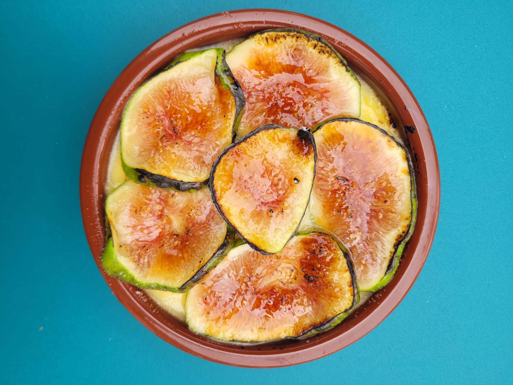 Sliced grilled sliced figs with caramelized edges, arranged in a terracotta bowl on a blue surface.