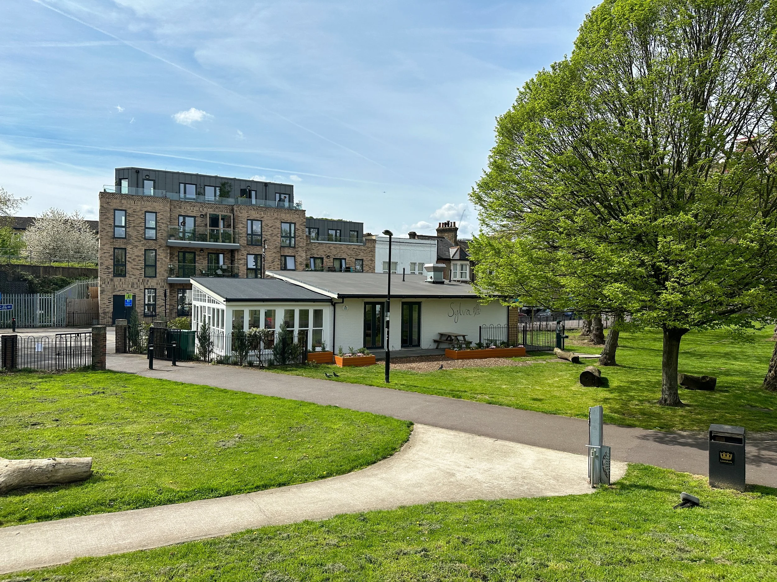 A park scene with green grass, a paved walking path, a large leafy green tree, and modern residential buildings in the background under a partly cloudy sky.