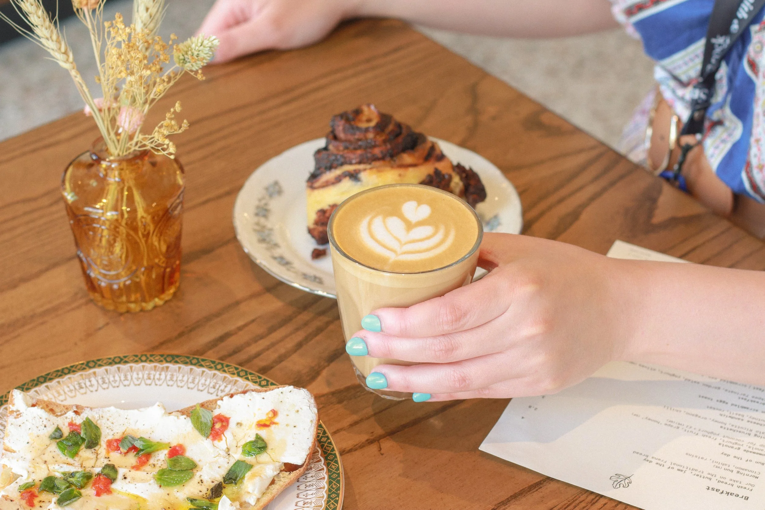 A hand holding a latte with latte art, a plate with bruschetta , a small vase with dried flowers, and part of a menu on a wooden table.