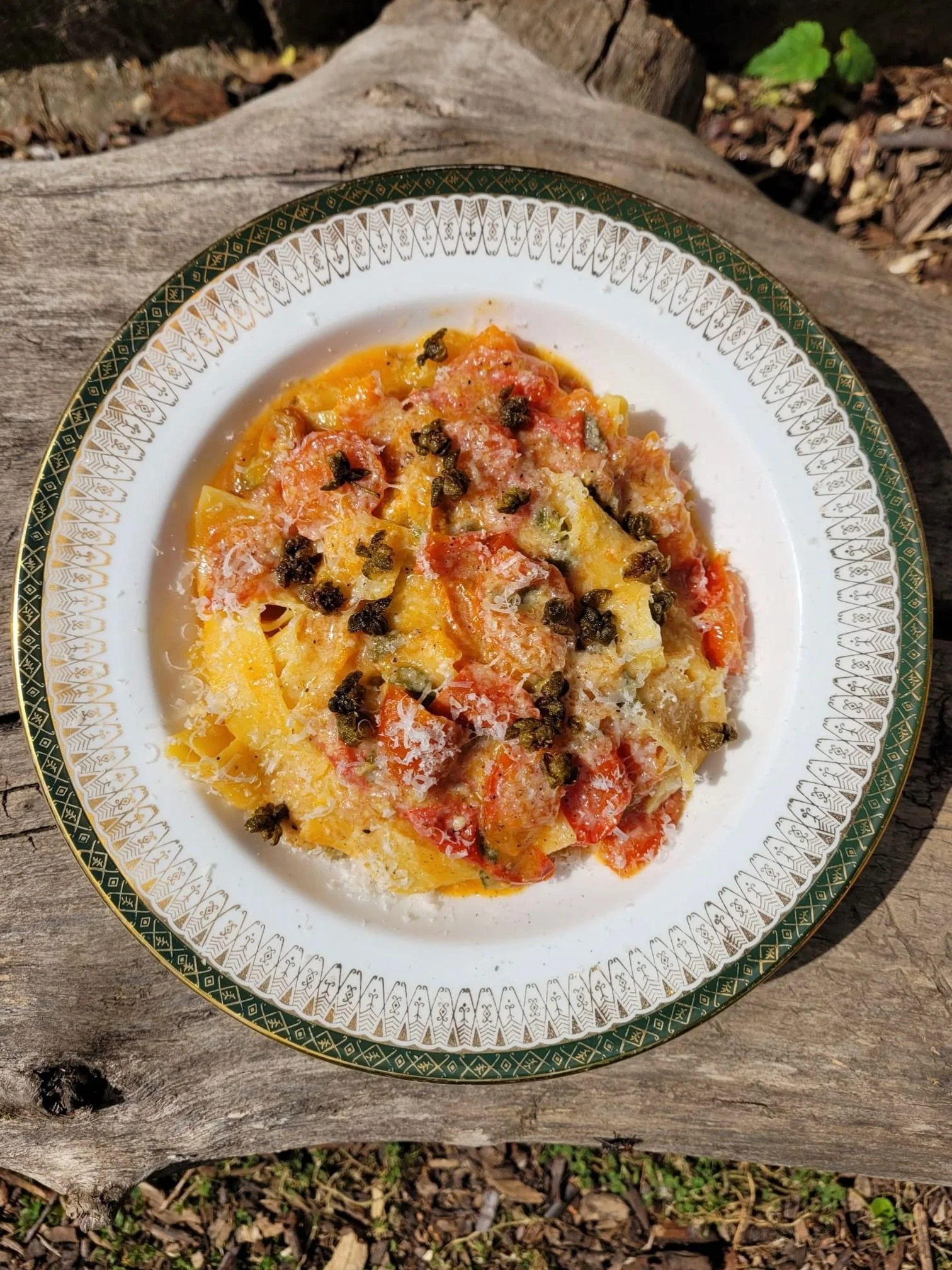 A plate of cooked pasta with tomato sauce, grated cheese, and black pepper, placed on a rustic wooden surface outdoors.