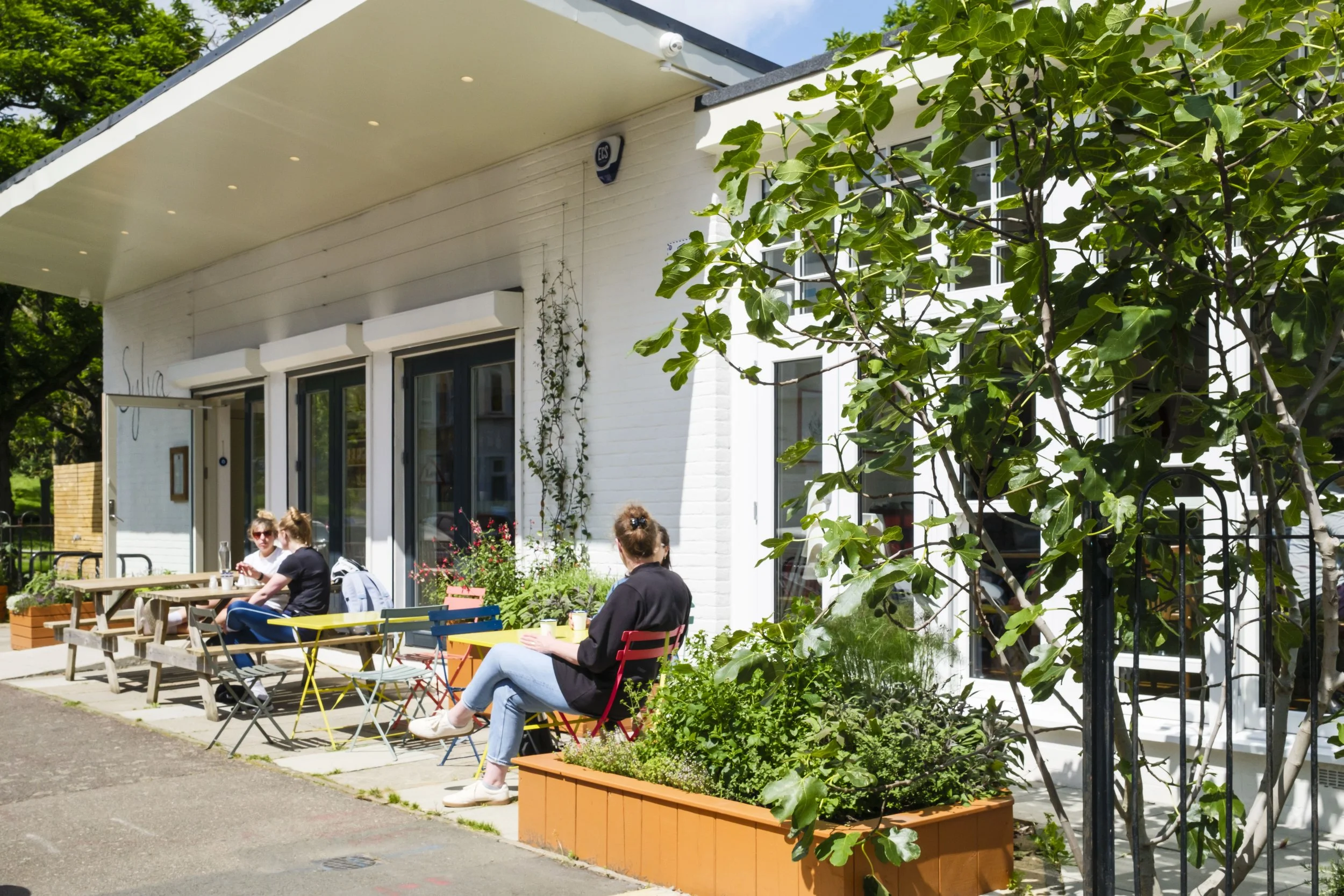 People sit outdoors at colorful tables and chairs in front of a white building with large windows and greenery, enjoying sunny weather.