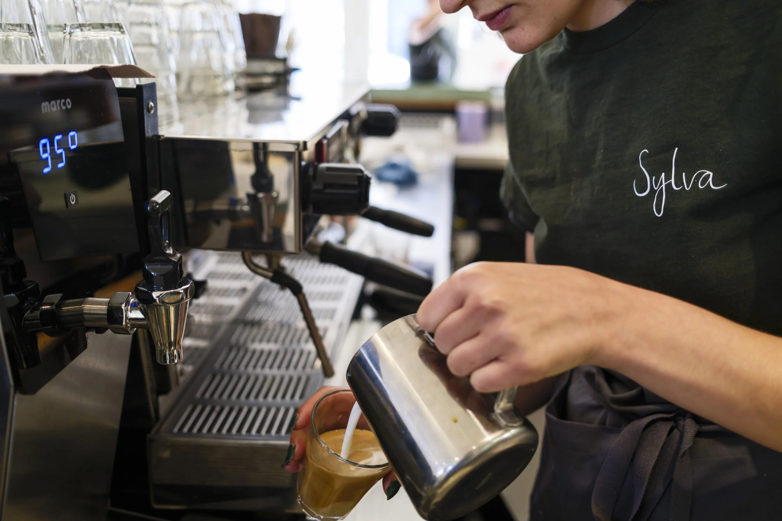 A barista pouring steamed milk into a coffee drink in a clear glass, with a professional espresso machine visible on the counter.