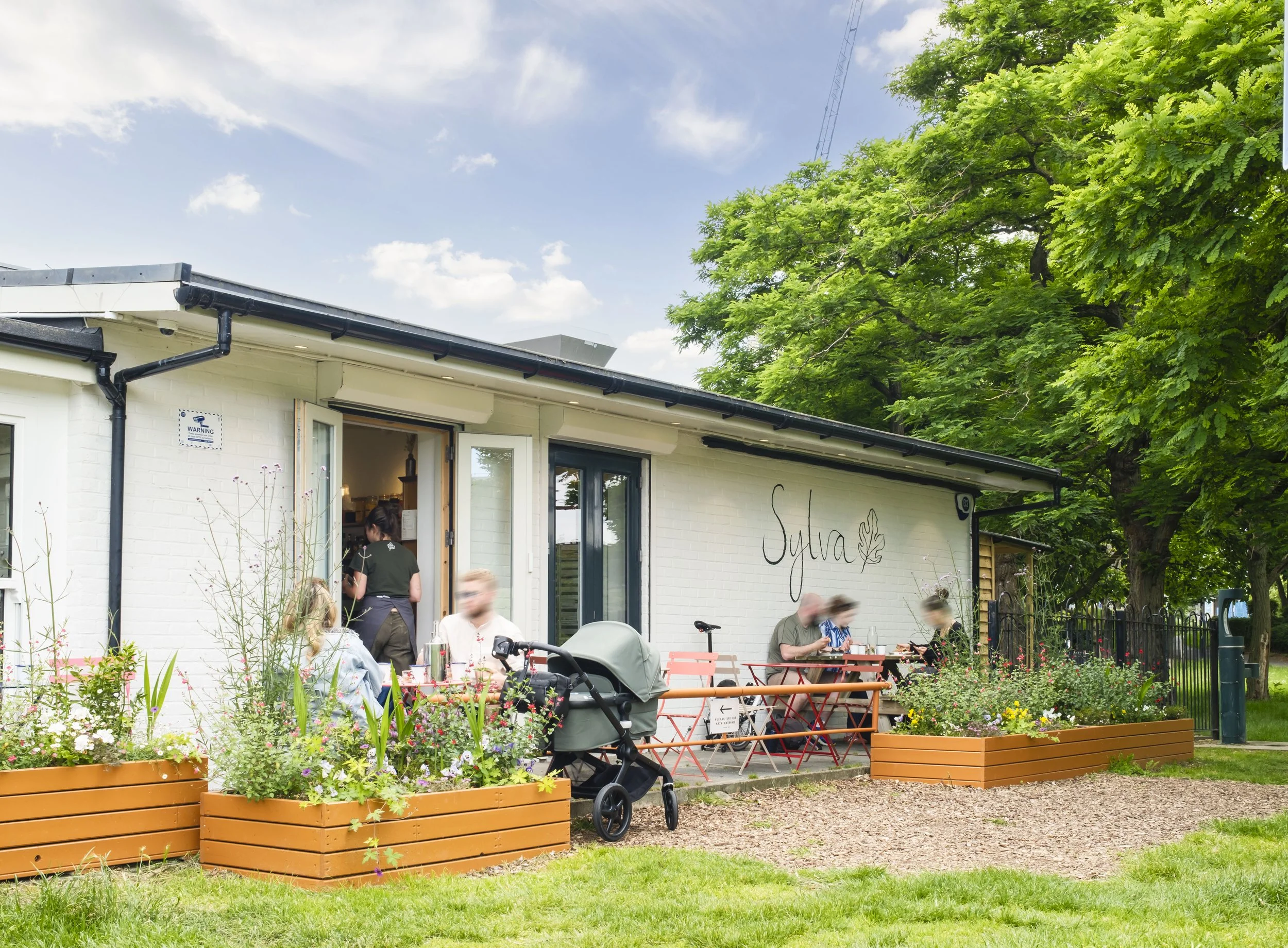 People dining outdoors at a restaurant cafe with orange planters filled with flowers in the foreground, a building with a sign that says 'Sylva,' large green trees, and a partly cloudy sky.