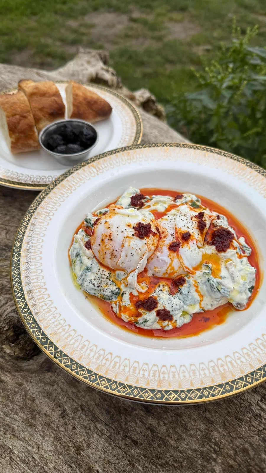 Poached eggs with spicy sauce on a decorative white and gold-rimmed plate, with bread and black olives on a separate dish in the background, outdoor setting with wooden surface and greenery.