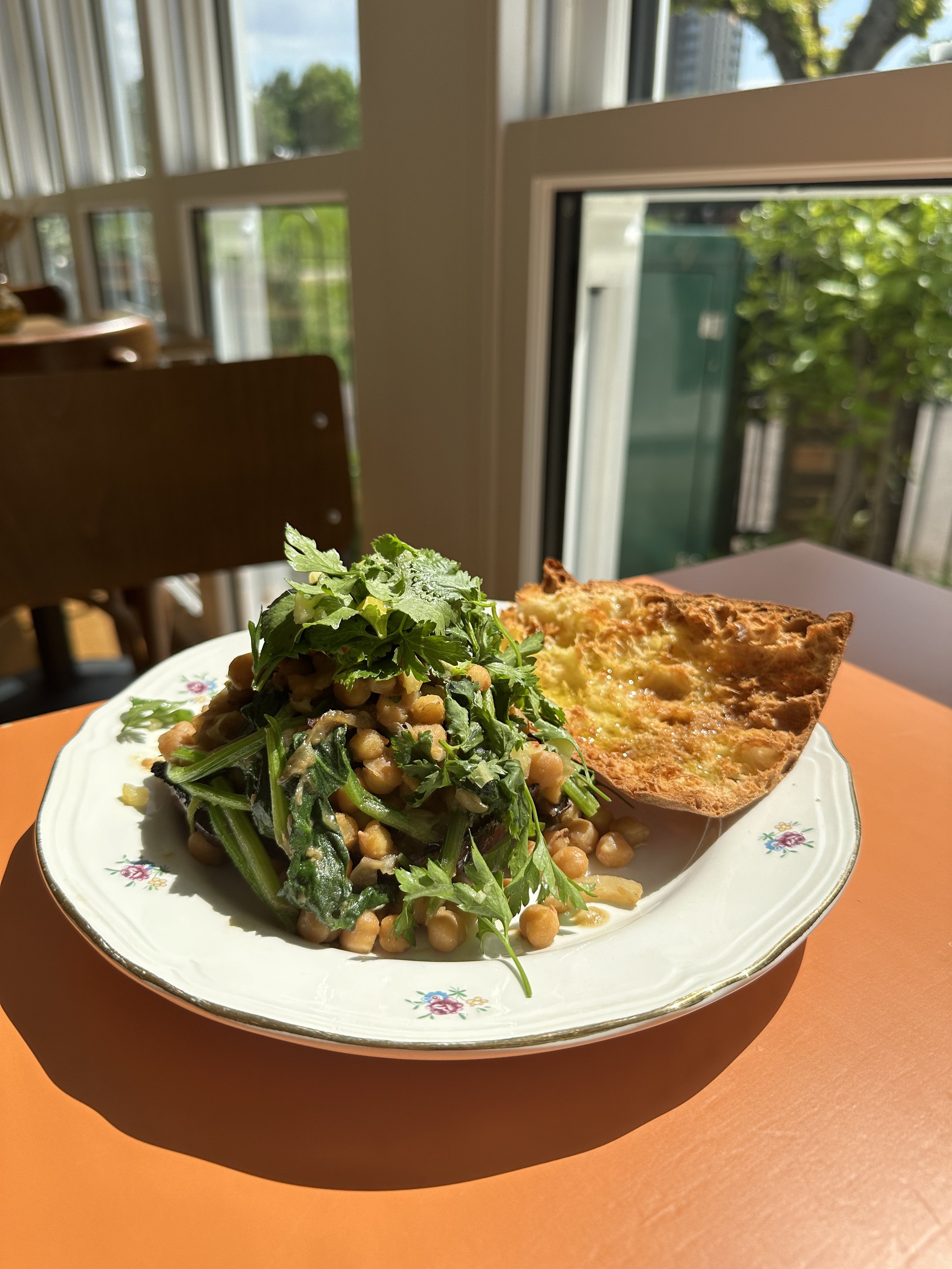 A plate with chickpeas, greens, and herbs, and a slice of toasted bread, placed on an orange table near a window with a view of trees and buildings outside.