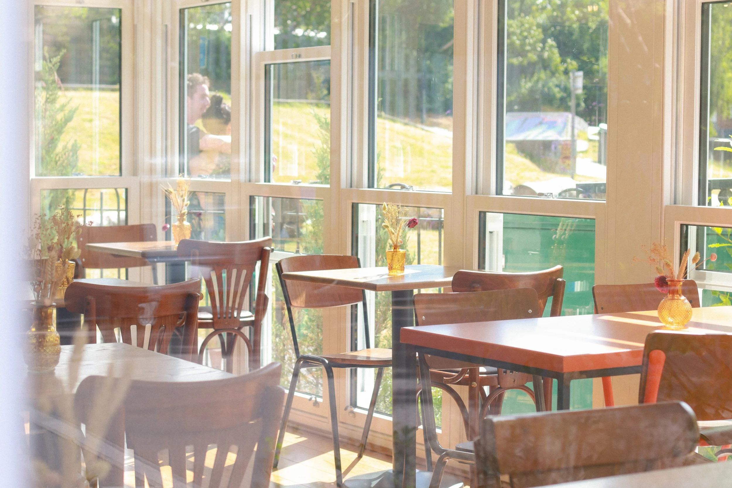 Sunlit interior of a cozy cafe or dining area with wooden tables and chairs, decorated with small vases and flowers, large windows showing green outdoor scenery.
