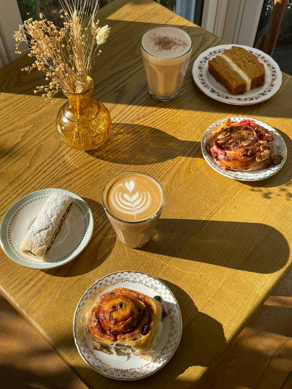 A wooden table with a vase of dried flowers, a latte with latte art, a glass of iced coffee with whipped topping, a piece of carrot cake, a almond bun, a cinnamon roll, and a pastry