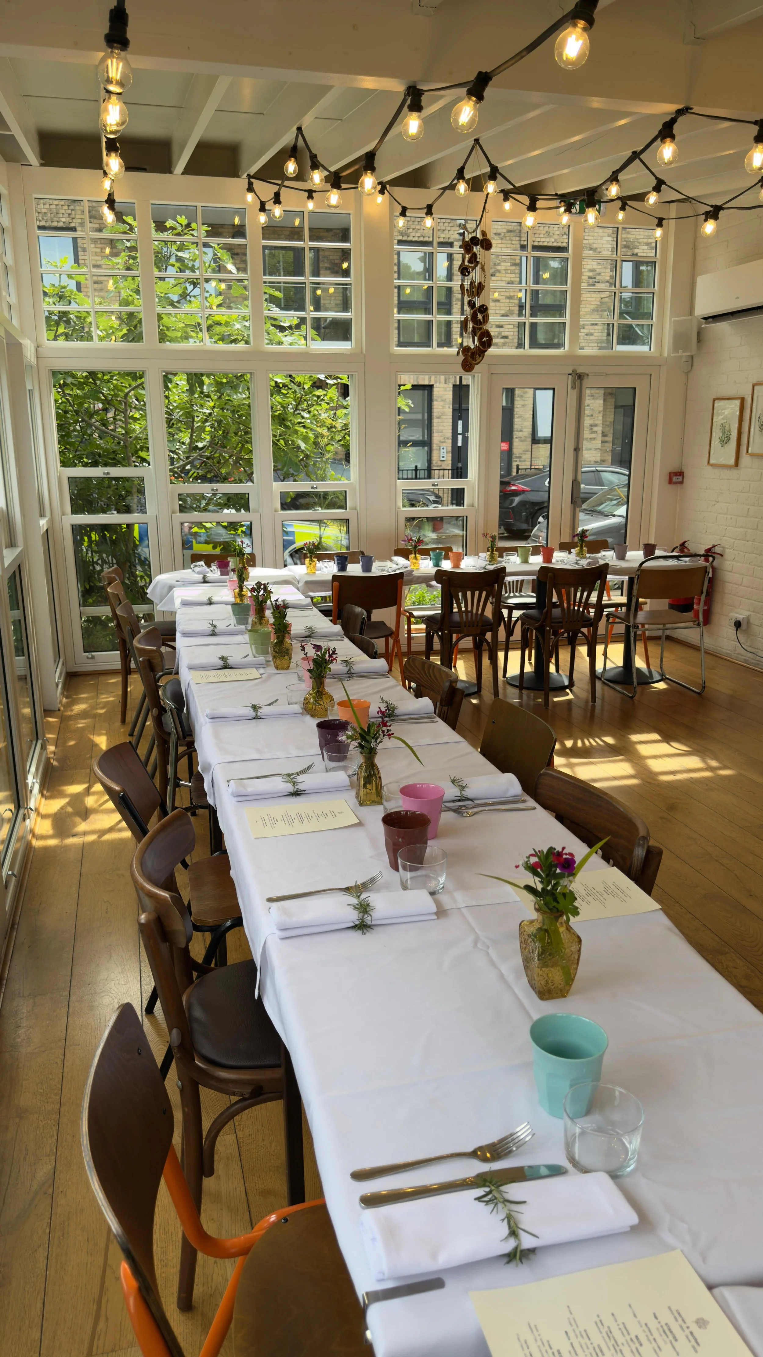 A long dining table set for a meal in a bright room with large windows and string lights.