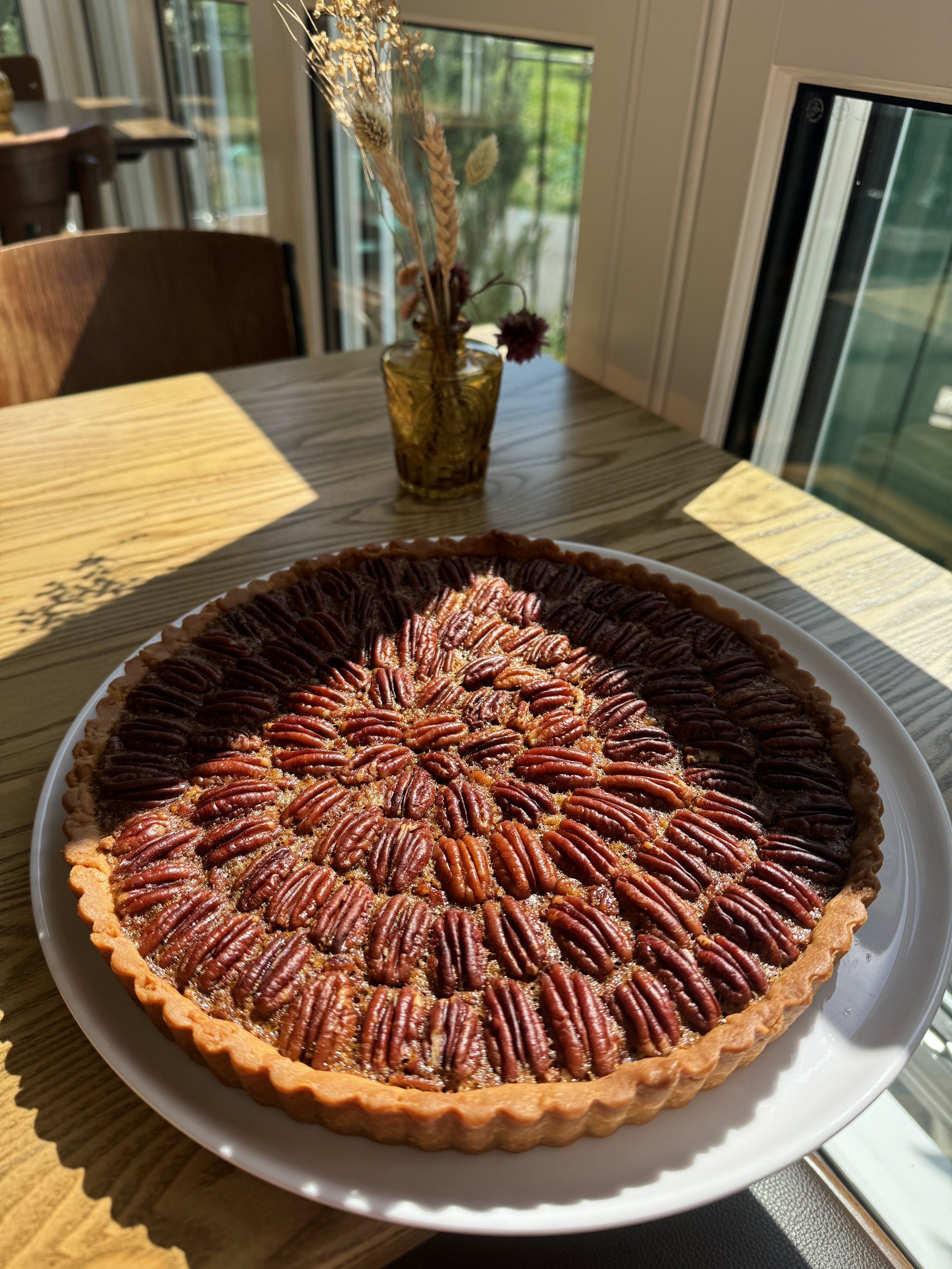 A pecan pie with a detailed topping of pecans on a white plate on a wooden table near a window with sunlight.