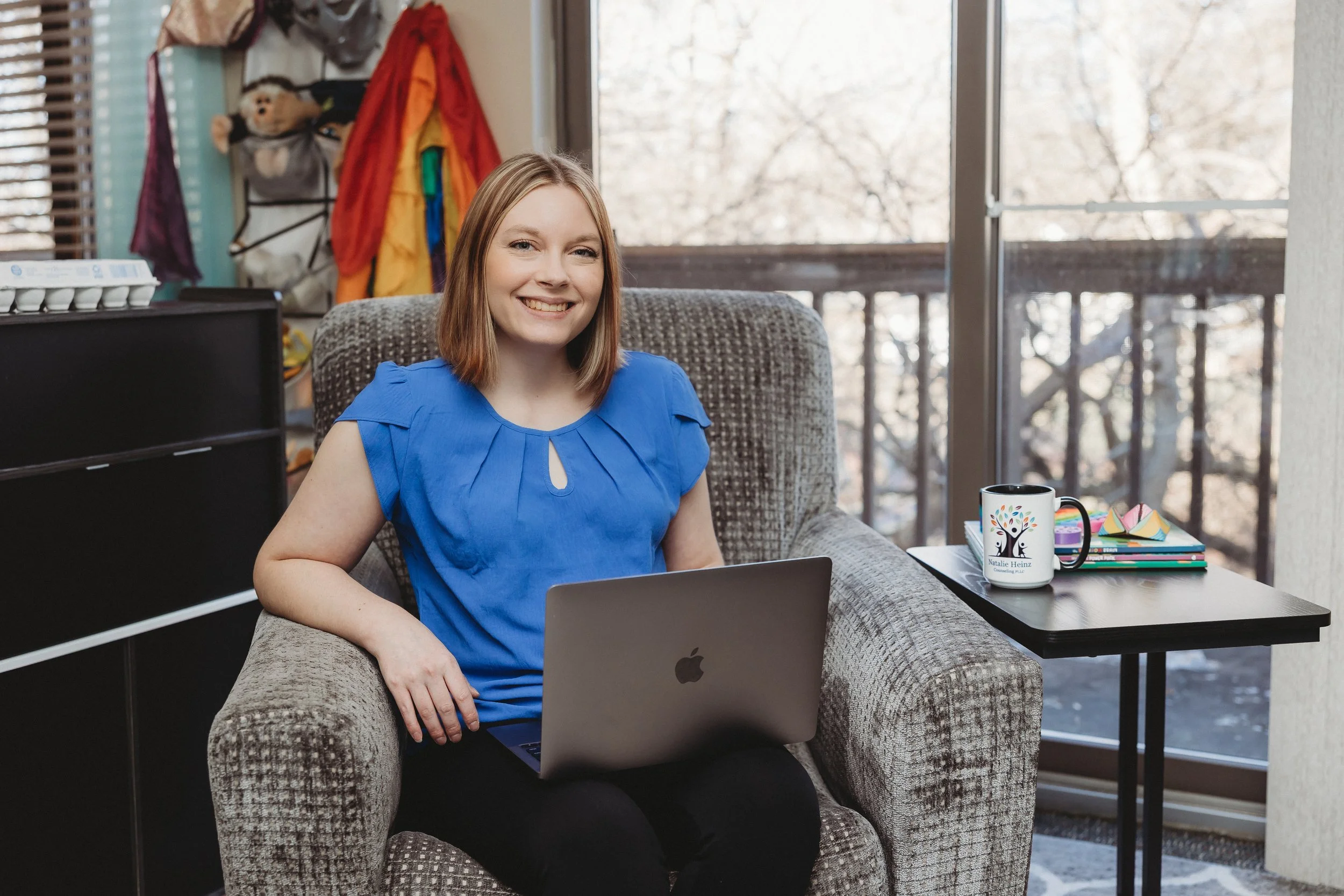 Natalie Heinz professional counselor seated and smiling in a blue shirt with a laptop sitting on her lap