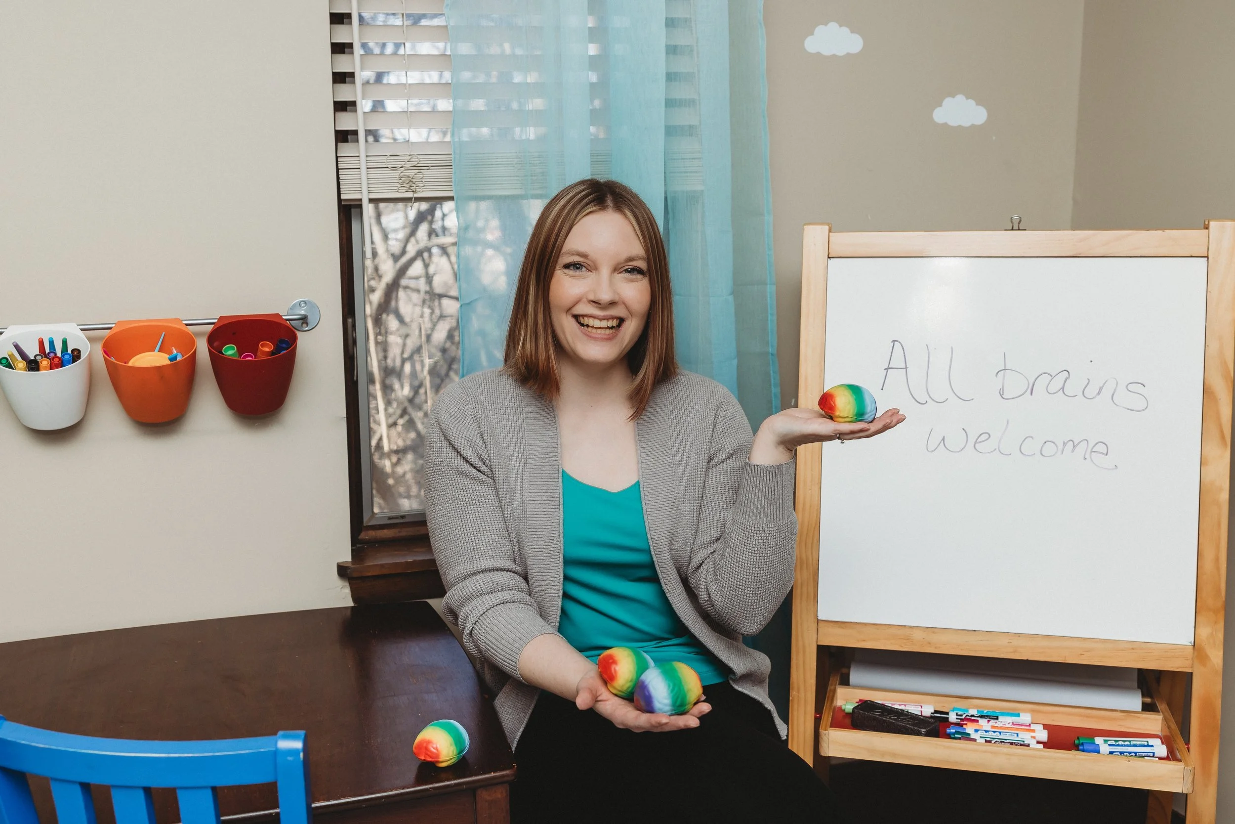 Natalie Heinz play therapist seated at a children's art table holding rainbow stress ball brains smiling with an easel in the background with the words All brains welcome.