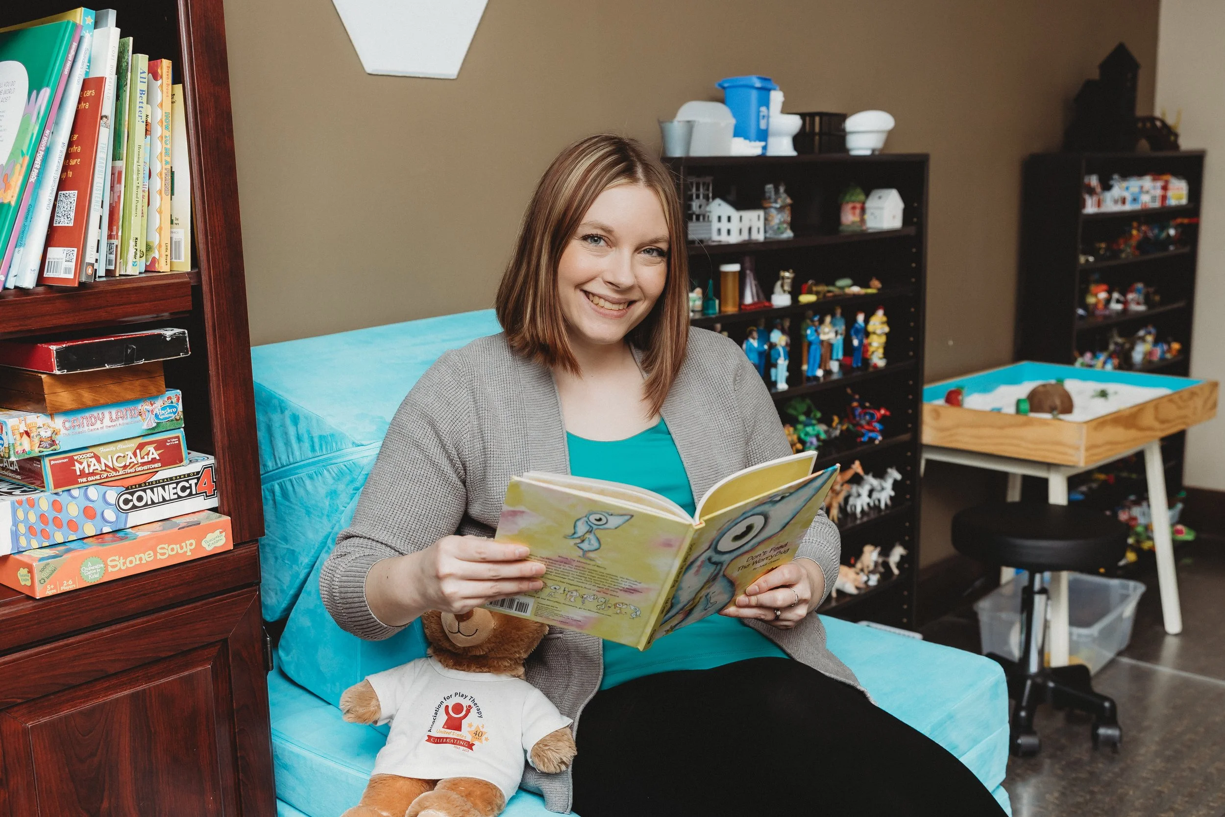Natalie Heinz counselor reading a children's book with a stuffed bear sitting on a light blue nugget couch with sand tray miniatures in the background