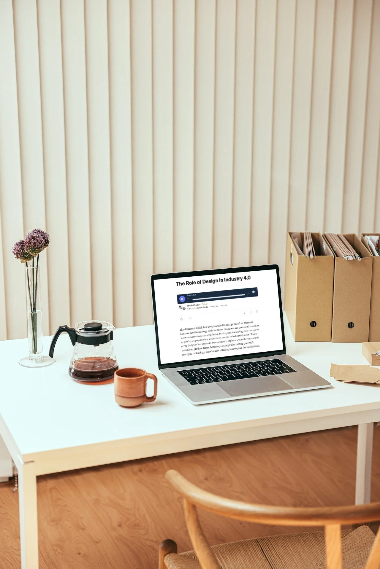 A white desk with a laptop, a glass coffee pot, a brown mug, a tall glass vase with purple flowers, and a cardboard organizer with files against a cream-colored striped wall.