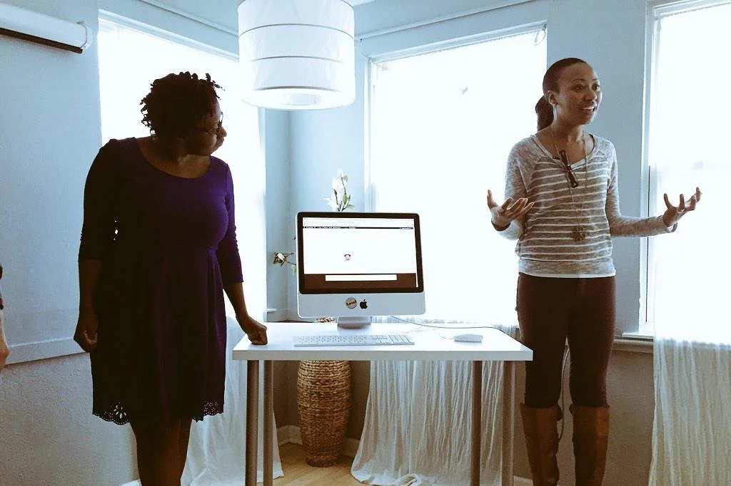 Two women having a discussion in a bright room with large windows, white walls, and a computer on a table between them.