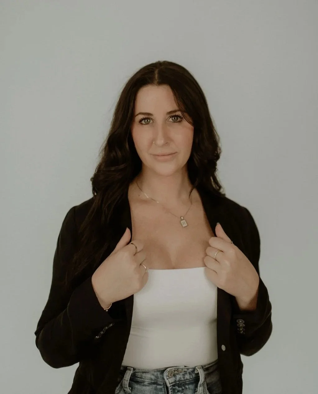Woman with dark hair wearing a black jacket and white top, posing with hands on lapels, and a necklace with a pendant.