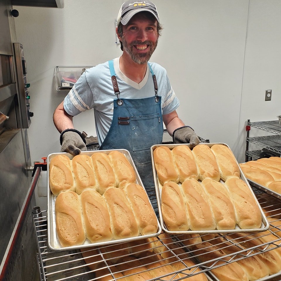 Man with a beard and gray cap smiling, holding two trays of freshly baked bread rolls in a bakery kitchen.
