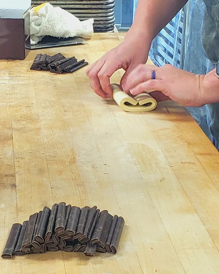A person is rolling out dough on a wooden surface, with chocolate curl cones arranged in a row in the foreground and chocolate curls and shredded white chocolate in the background.