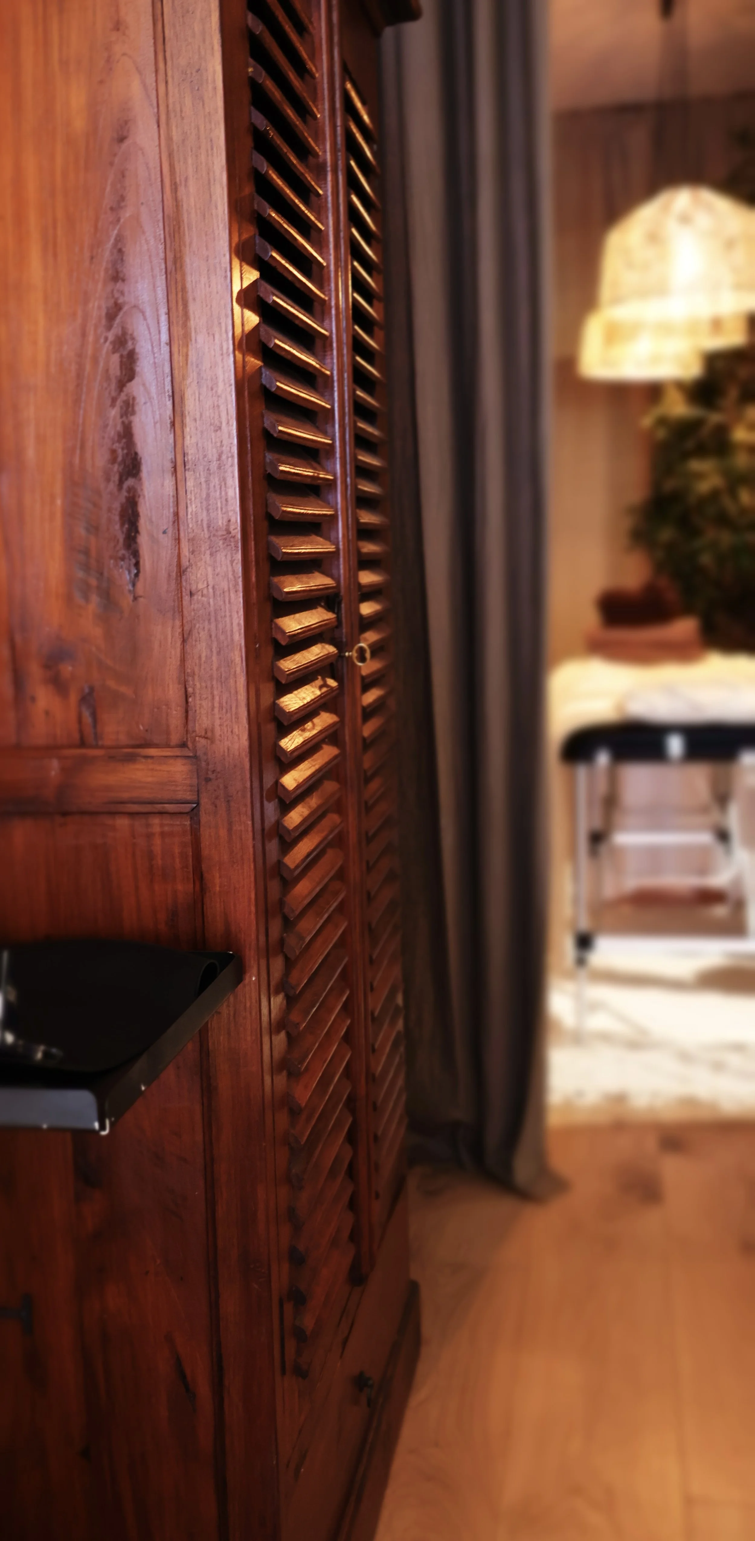 Wooden wardrobe with louvered doors, a small shelf, and a blurred view of a cozy room setting with a table and lamp.