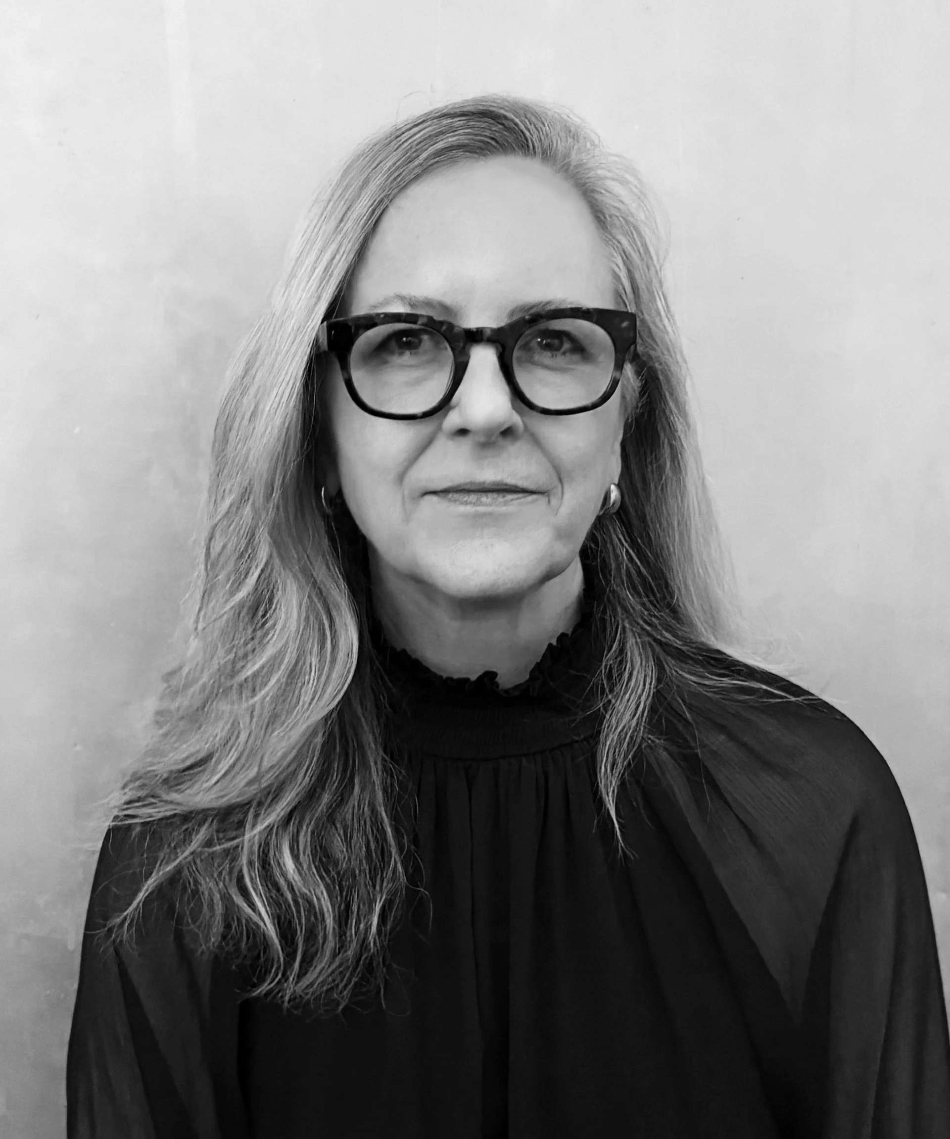 A black-and-white portrait of a woman with long, wavy hair and glasses. She is wearing a dark top and is standing against a plain, light-colored background.
