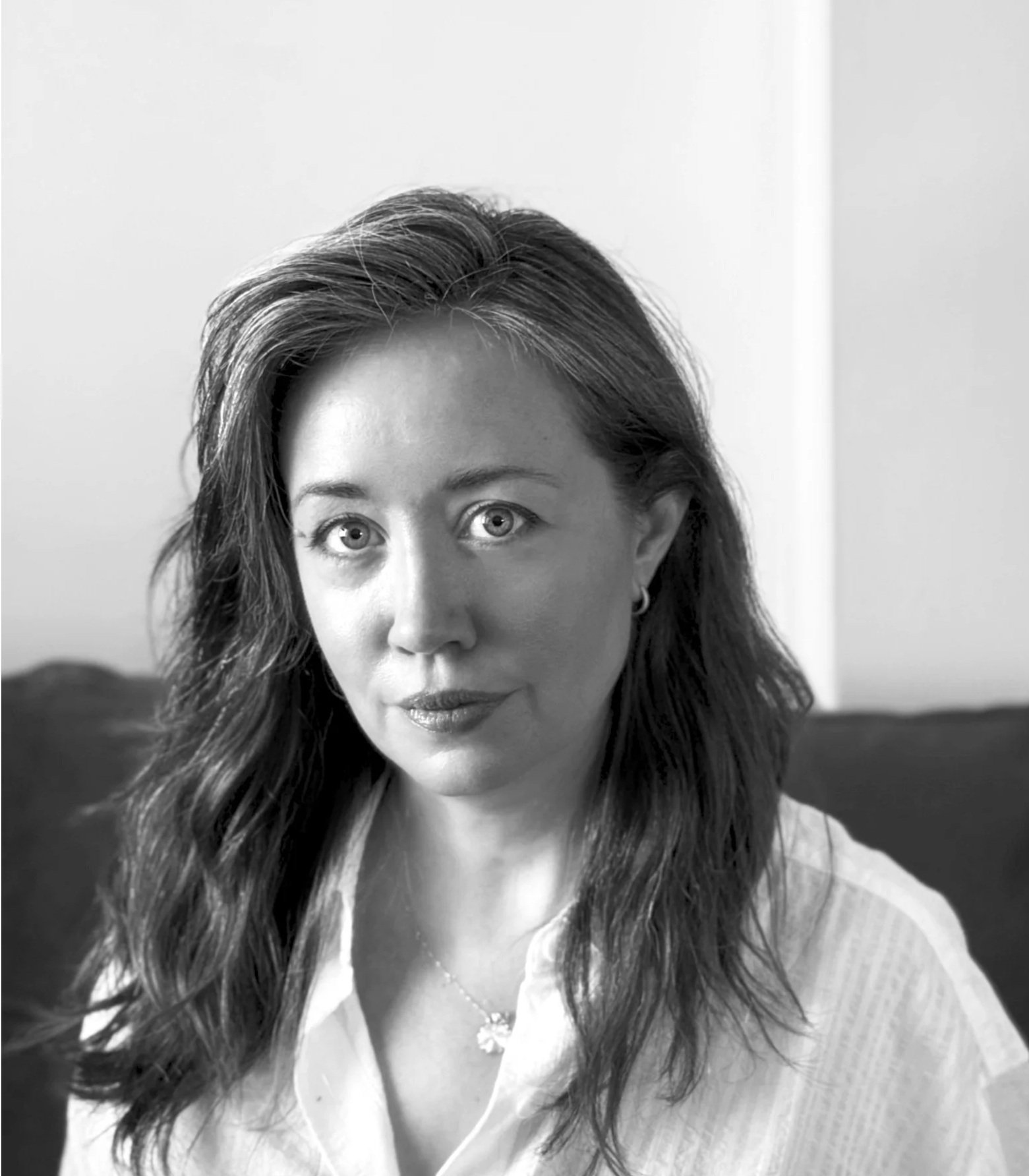 A black and white photo of a woman with long wavy hair, wearing a white top and a necklace, looking directly at the camera.