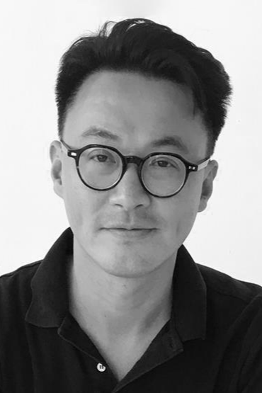 A black-and-white photo of a man with short dark hair, wearing round glasses and a black collared shirt, smiling gently at the camera.
