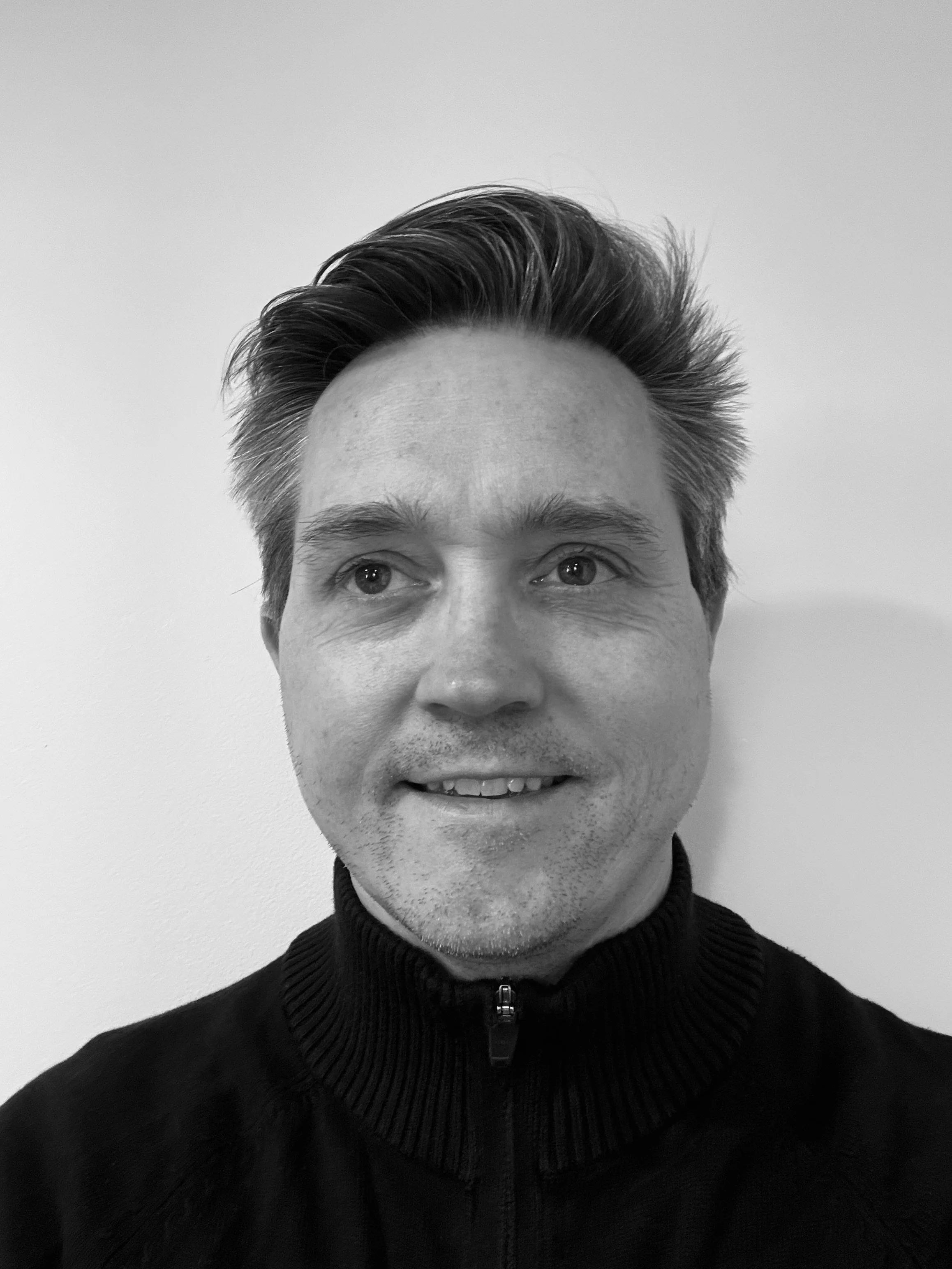 Black and white photo of a man with styled hair, light skin, smiling slightly, wearing a black zip-up sweater with a ribbed collar, against a plain light-colored background.