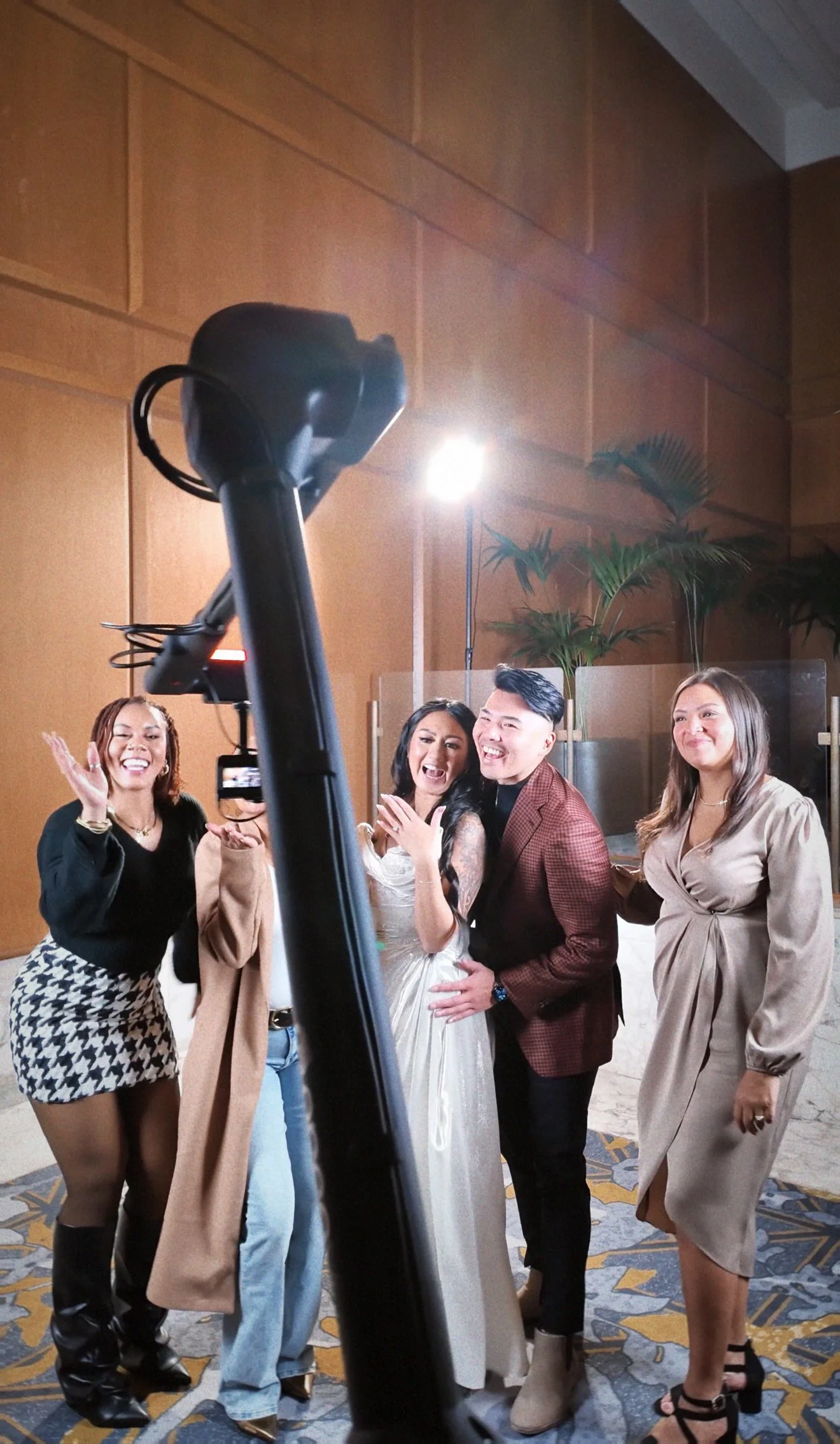 Group of five people taking a selfie at a social event, with a camera and tripod visible in the foreground. The group is diverse, smiling, and dressed in stylish attire, with a wooden-paneled wall and some plants in the background.