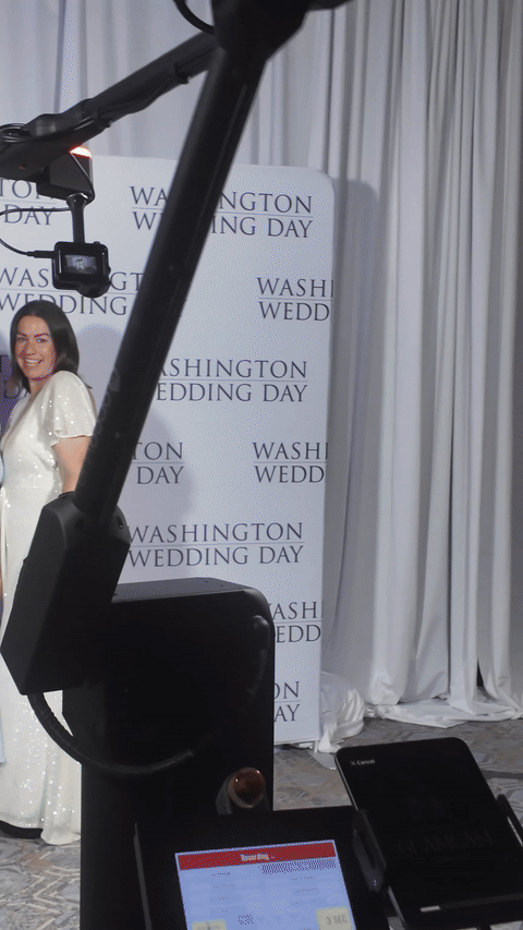 Partial view of a woman in a white dress at a wedding event, with a photo backdrop reading 'Washington Wedding Day' and some camera equipment in the foreground.