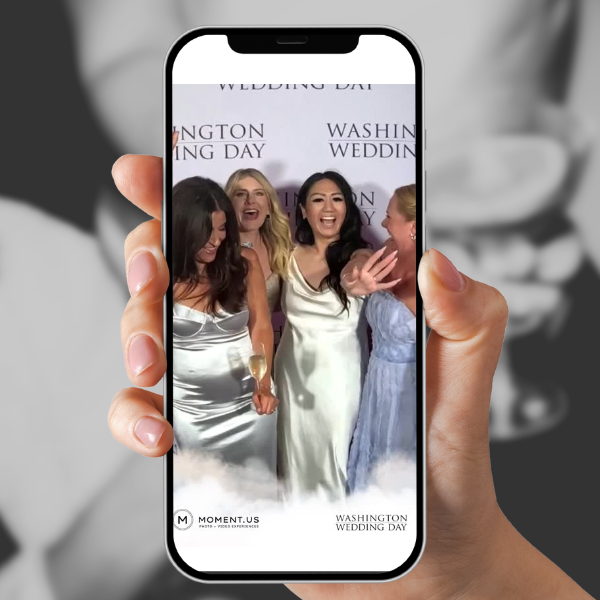 A person holding a smartphone captures a photo of four women dressed for a wedding, smiling and laughing together in front of a step-and-repeat backdrop with text related to the wedding day.