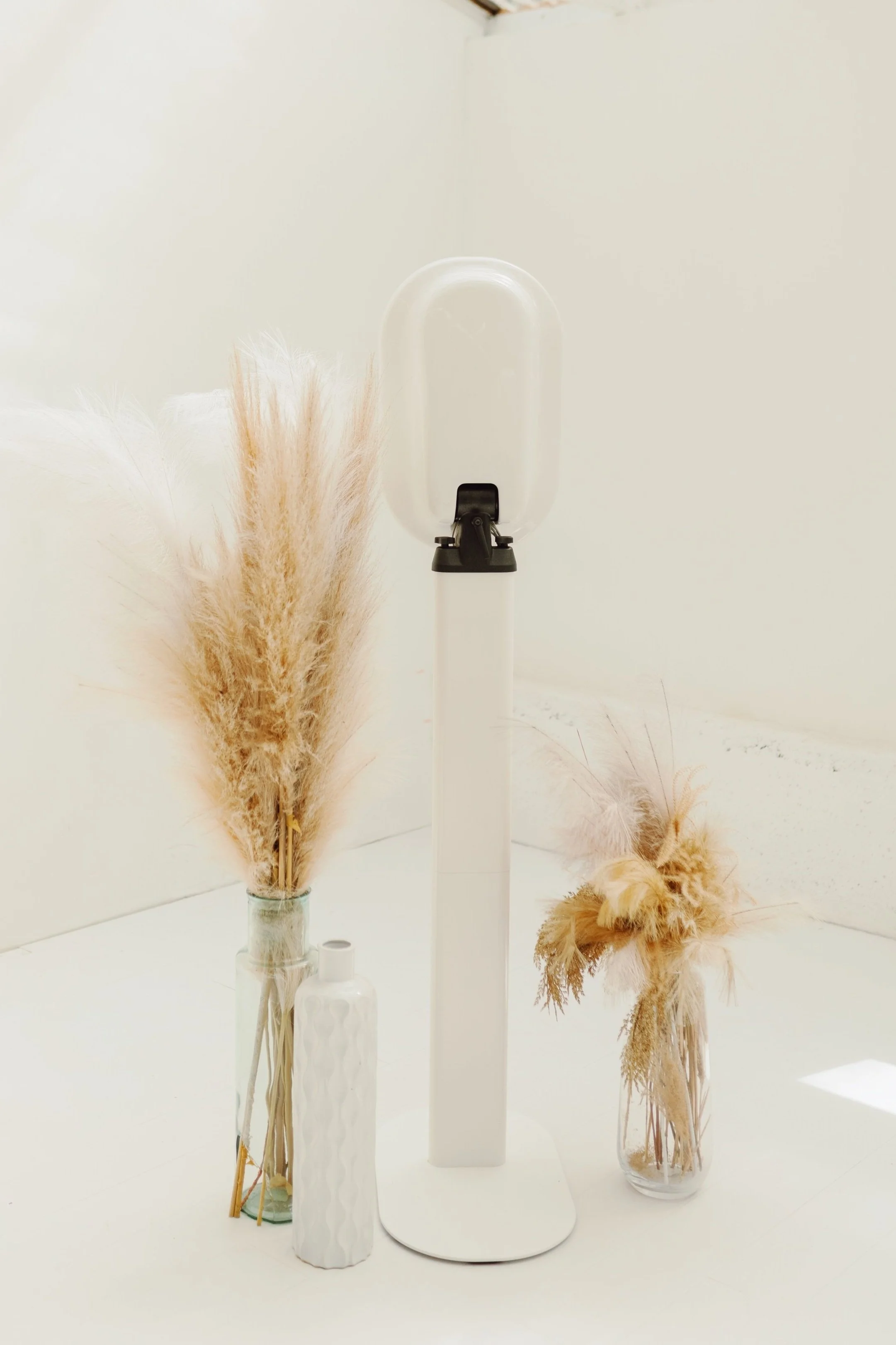 Decorative neutral-colored dried flower arrangements in vases and a sleek white lampshade on a minimalist white table.
