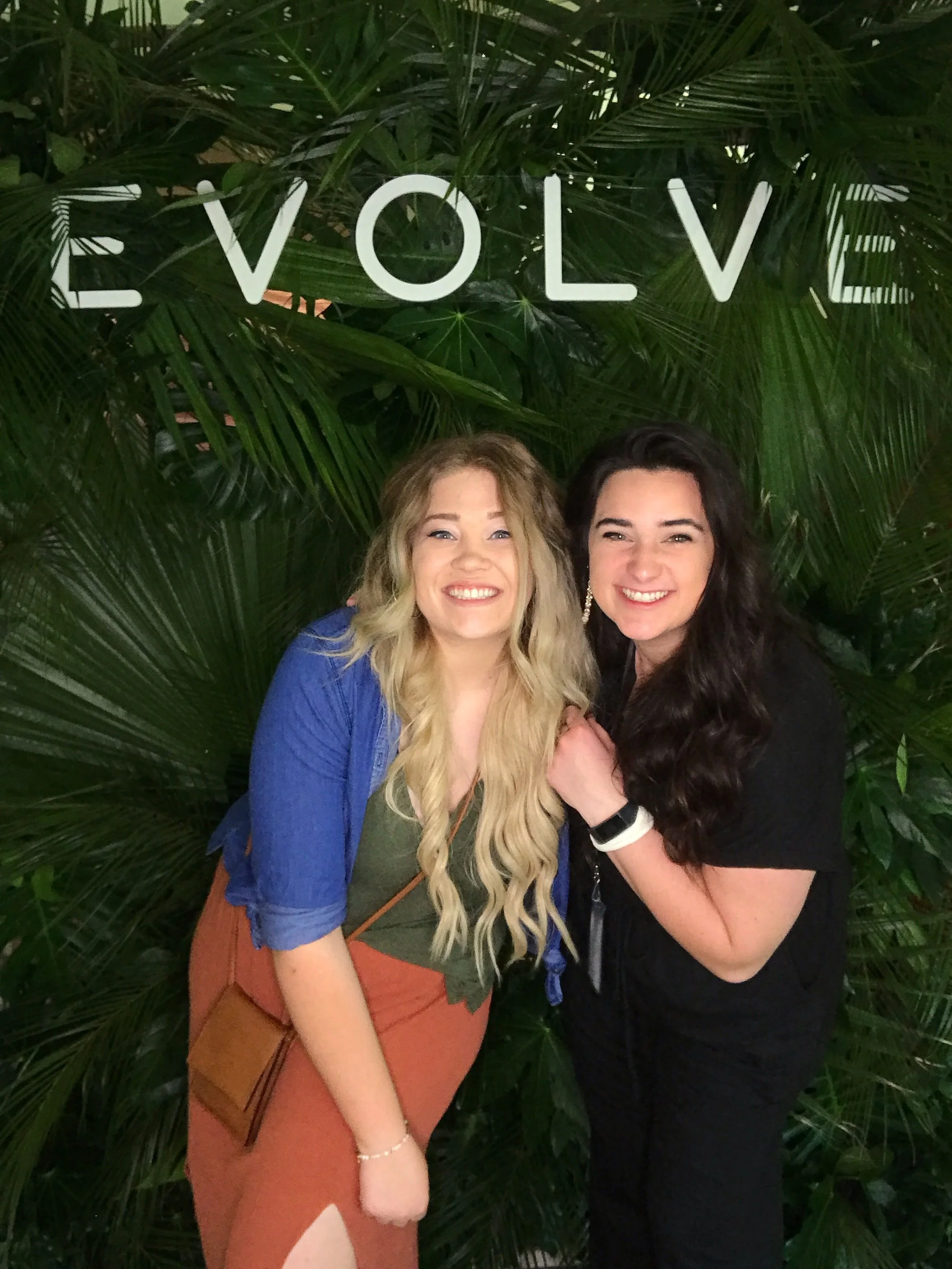 Two women smiling in front of green tropical plants with the word 'Evolve' displayed above them.