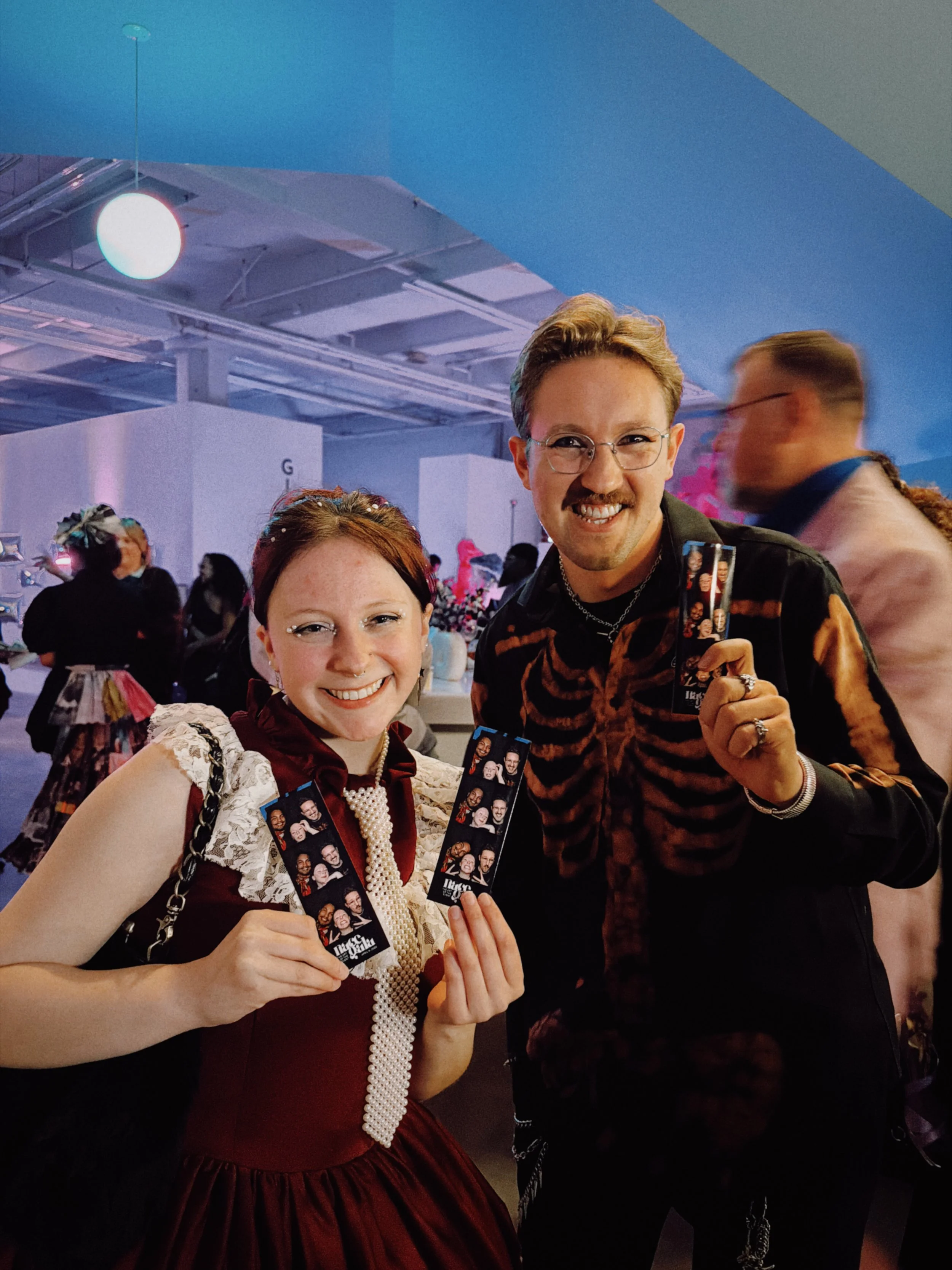 Two smiling people holding photo booth strips at a lively indoor event, with other attendees in the background.