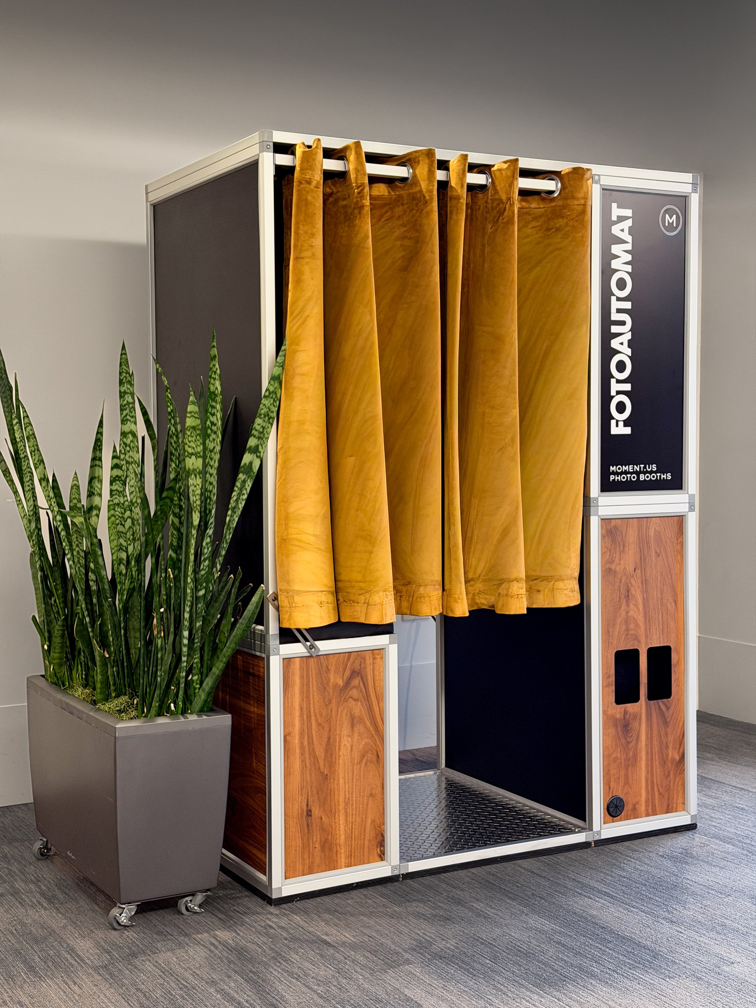 A photo booth with brown curtains and a black interior, labeled "FOOTOMAT" with a sign indicating it's a photo booth company, and a tall green plant in a gray rectangular pot on wheels in front of it, set on a gray carpeted floor against a plain wall.