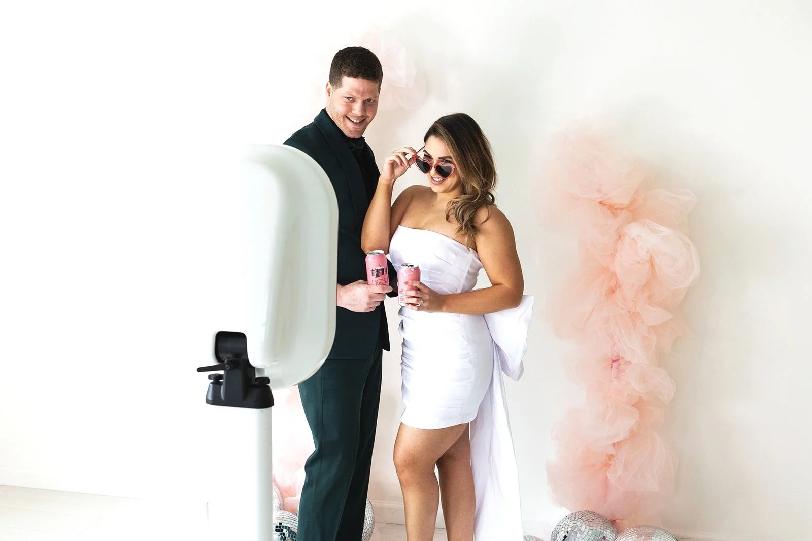 A man and woman, dressed for a celebration, posing for a photo in front of pink tulle decorations and disco balls, holding cans of beverage, with the woman adjusting her sunglasses.