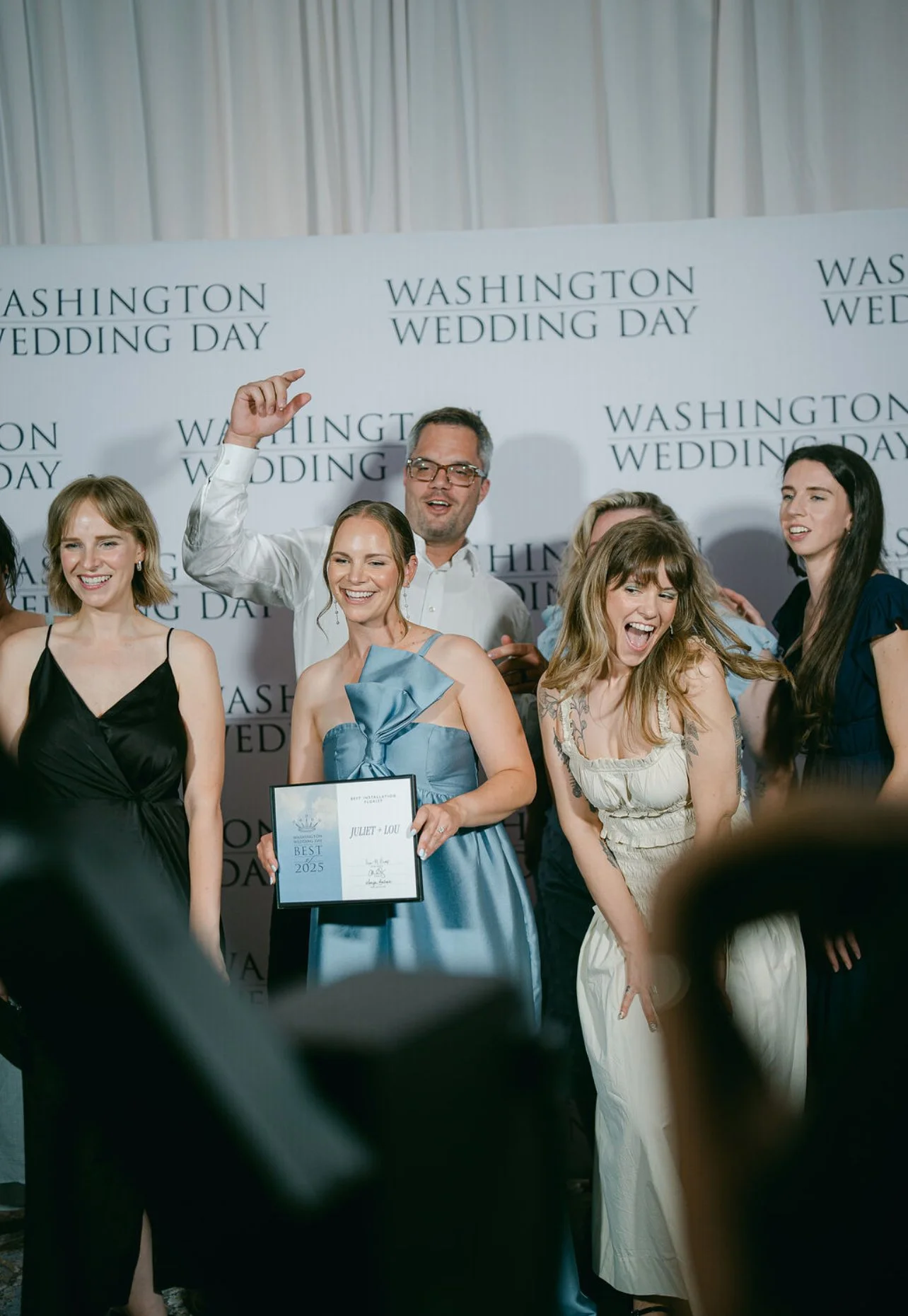 Group of people celebrating at a wedding event, with a banner reading 'Washington Wedding Day' in the background. One woman holds a certificate for best wedding couple 2025, smiling and surrounded by friends.